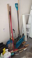 Multiple long-handled gardening tools including shovel, broom, snow shovel, rake, some small hand tools, rubber boots, and watering can grouped on a floor corner.