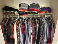 Wide view of men's shirts hung on hangers along closet rod with folded shorts or t-shirts stacked on shelf above