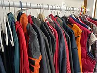 Photo showing various men's jackets and vests hanging on a rack, including puffer styles in red, orange, and blue, fleece jackets, and plaid shirts.