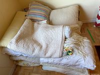 Stack of pillows and quilts on floor showing different textures, fringe, and patterns.