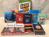 Image showing 11 card games and playing card decks arranged on a white cloth backdrop. Includes Skip-Bo, Three Seas, Memory Game, Mindful Mix, Happy Confident Me Affirmations, Western Genius, two decks of Queeng Playing Fair, Canada 45 Different Images, and another deck with a green Ltd. logo.