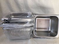 Four new stainless steel commercial food pans stacked, three wrapped in plastic, one exposed showing clean interior.