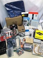 Wide view showing many boxed and bagged electronics accessories including Cisco box, OtterBox case, cables, USB hubs, and mounting bracket.
