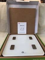Open box displaying the white 1 By One smart scale with four metal sensors and instruction manual.