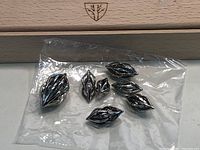 Seven metal dice with intricate hollow leaf designs inside plastic packaging, placed on top of a wooden box lid with emblem.