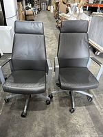 Front view of two black leather executive conference chairs with high backs and metal armrests on casters placed side-by-side on warehouse floor.