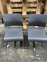 Two armless side chairs front view showing grey seats and chrome legs on concrete floor with wooden pallet background.