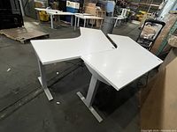 Photo of two white boomerang-shaped desks next to each other in a warehouse setting, showing tops and bases with metal legs.