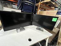 Two Dell 24-inch monitors side by side on a white surface with shelves and boxes in the background.