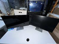 Two Dell monitors side by side on a white table against storage boxes in background. Screens are black and reflective with Dell logos on bottom bezel.