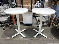 Two white hightop oval tables with metal bases, shown side by side from the front on a warehouse floor.