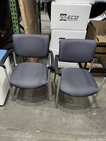 Two gray office side chairs with metal legs and fixed black armrests photographed from front on concrete floor