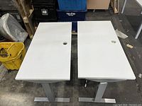 Two white variable height riser desks side by side with visible drilled holes on the surfaces and minor scratches on the legs and tabletops.