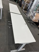 Photo showing two white desks with smooth surfaces and T-shaped legs in a warehouse. Minor scratches visible. Cable management holes on top.