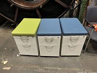 Photo of three white rolling file cabinets with cushioned tops in green and blue, arranged side by side on a concrete floor.