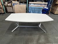 White large conference table with trapezoidal curved top sitting on metal base in warehouse environment taken from side angle.