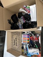 Box containing various office supplies including markers, pens, stapler, tape dispenser, scissors, clips, and other small items.