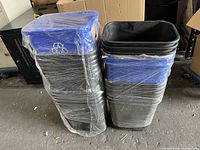 Stacked black plastic trash bins with blue recycle lids, wrapped in plastic, approximately 40 units.