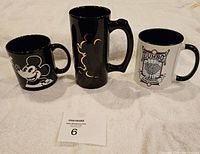 Photo showing the front side of three Disney-themed mugs: black Mickey Mouse face mug, tall black Mickey Mouse silhouette mug, and white Dumbo mug.
