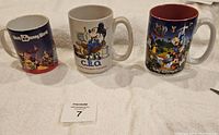 Three Disney coffee mugs with different character and park-themed prints placed on a white towel.