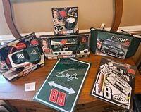 Overview of the lot showing boxed die-cast cars, metal sign, notebook with pen, and a lunch bag all themed around Dale Earnhardt Jr. NASCAR memorabilia.