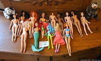Photo showing 12 dolls laid out on a wooden surface, including Barbie-style dolls and two mermaid dolls, some missing clothing or heads.