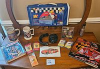 Wide view showing all items in the lot including two NASCAR mugs, booklets, NASCAR trading cards, headphones, a printable car image, and a NASCAR collectible bag with a handle.