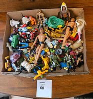 A top view of a cardboard box filled with assorted action figures and toys. Includes humanoid figures, animal figurines, and toy vehicles, all placed in a pile.