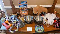 Photo showing the two stainless steel bowls, various rope and bone-shaped chew toys, dog clothing, glow-in-the-dark collar packaging, and PediPaws nail trimmer box all arranged on a wooden bench