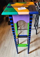 Front view of side table showing colorful pop art top, striped legs, and purple apron.