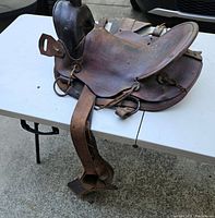 Vintage leather horse saddle placed on a white folding table, showing full side view with attached stirrup and straps.