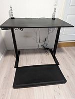 Full view of black Evodesk sit-stand desk with two transparent speakers on top and black standing mat in front.