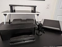 Full set of home office accessories displayed on a black surface against a white wall; includes foot rest, microphone, cable storage box, hole punch, monitor stand, laptop stand and tray desk