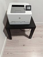 Front view of white HP Color LaserJet M255dw printer on black IKEA LACK table against a white wall, displaying touchscreen and paper tray top cover.