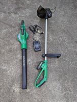 Full set of green Certified lawn care cordless tools including leaf blower, string trimmer, battery, and charger on concrete surface.