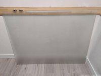 Frosted quarter glass whiteboard with clear edges leaning against a white wall on wood flooring. Top edge has metal wall mounts attached.