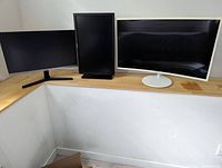 Three monitors on a wooden desk; the leftmost is Samsung 24" LED-lit monitor, middle is Dell 24" monitor displayed vertically, rightmost is white curved Samsung 34" monitor.