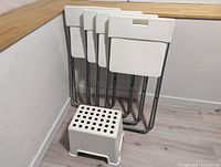 Four white Ikea GUNDE folding chairs stacked against a wall with the white footstool in front.