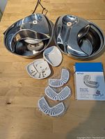 Two stainless steel Pioneer Pet fountains in raindrop design with white plastic pump housings, dishes, and three unopened replacement filters on a wooden table.