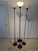 Full view of three standing black torchiere floor lamps together, showing bases and cords on floor.