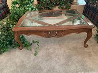 Front-angle view of a wooden framed coffee table with geometric glass top and carved details, placed on carpeted floor near plants and sofa.