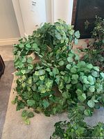 Four artificial plants with green foliage placed on a carpeted floor. Plants have dense leafy vines and leaves in black plastic pots.