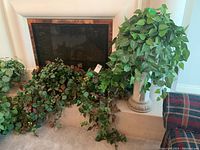 Photo showing three artificial plants around a fireplace, highlighting the tall plant in a ceramic stand and two trailing vine plants with green and reddish leaves.