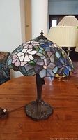 Full view of the stained glass style lamp on a wood table, showing the colorful floral patterned plastic shade and bronze metal base.