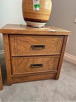 Front view of wooden nightstand showing two drawers with metal handles and wood grain finish, with lamp and cord on top surface.