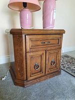 Angled side view of wood nightstand showing carved side detail, drawer and two doors with metal ring pulls, wooden top surface, and some scratches on base.