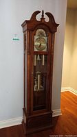 Full front view of Ridgeway grandfather clock placed against wall showing ornate bonnet top and brass clock face.