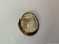 Front view of the oval shell cameo brooch showing the detailed carving of the scene with the woman, door, wall, trees, and houses framed in yellow gold-filled bezel setting.