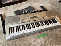 Full top view of Casio keyboard on carpet with disassembled black keyboard stand parts and metal music book stand beside it.