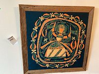 Photo showing framed square silkscreen art painting with a stylized figure and floral border on a dark background, mounted in a wooden frame.